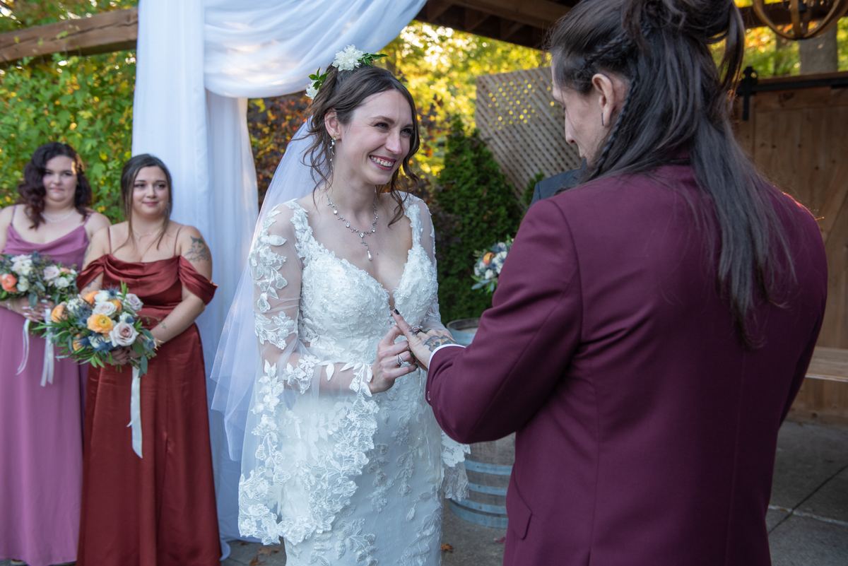 A bride in a lace gown smiles as she exchanges rings with her partner under draped fabric and trees.