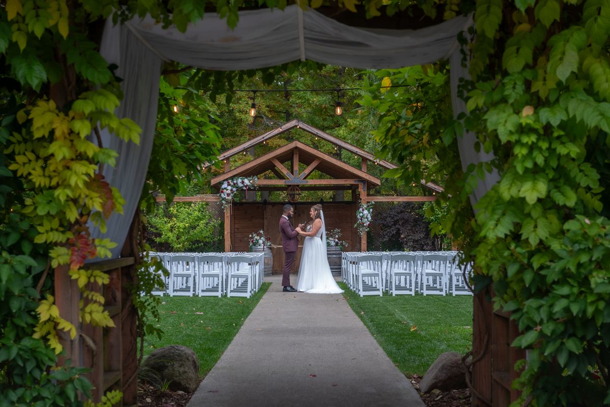 Alexandria and Derado stand at the end of a paved aisle beneath a wooden arbor, framed by lush greenery and draped fabric.