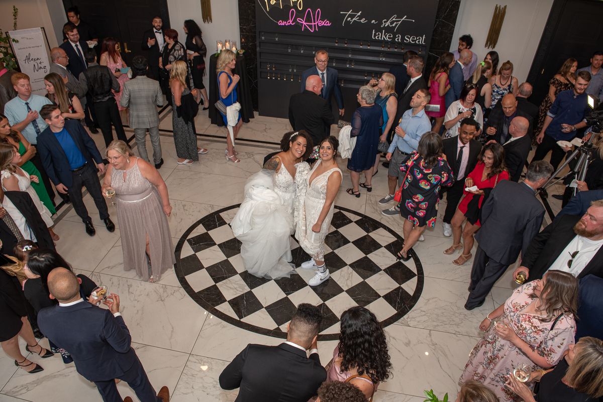 Two brides stand on a black-and-white checkered floor at a warmly lit wedding reception with guests mingling.