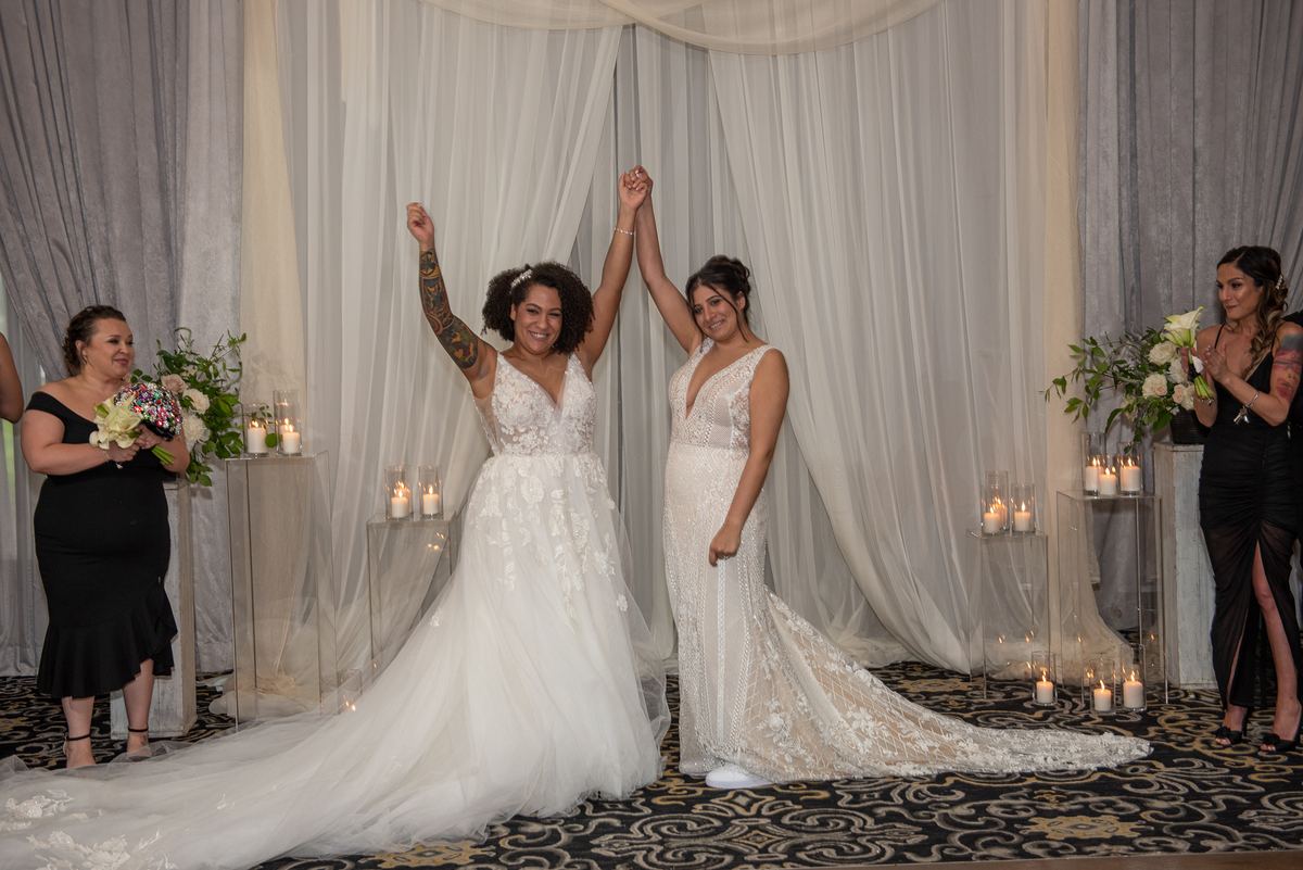 Two brides raise their hands in triumph, smiling joyfully at their wedding ceremony surrounded by candlelight.