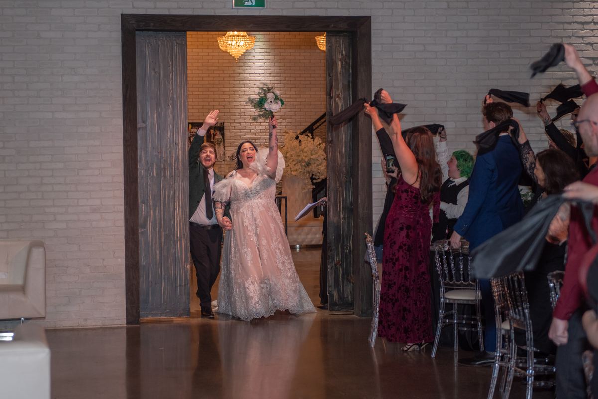 Bride and groom burst through wooden doorway into reception hall, guests throwing black fabric in celebration.