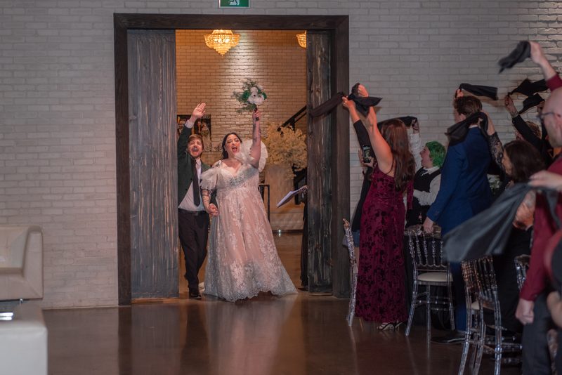 Bride and groom burst through wooden doorway into reception hall, guests throwing black fabric in celebration.