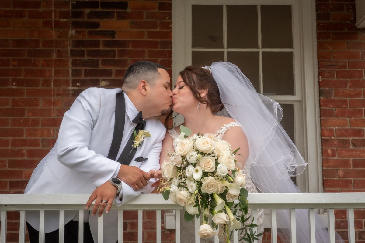 A bride in a white veil and bouquet kisses her groom in a tuxedo on a balcony, illuminated by soft daylight.
