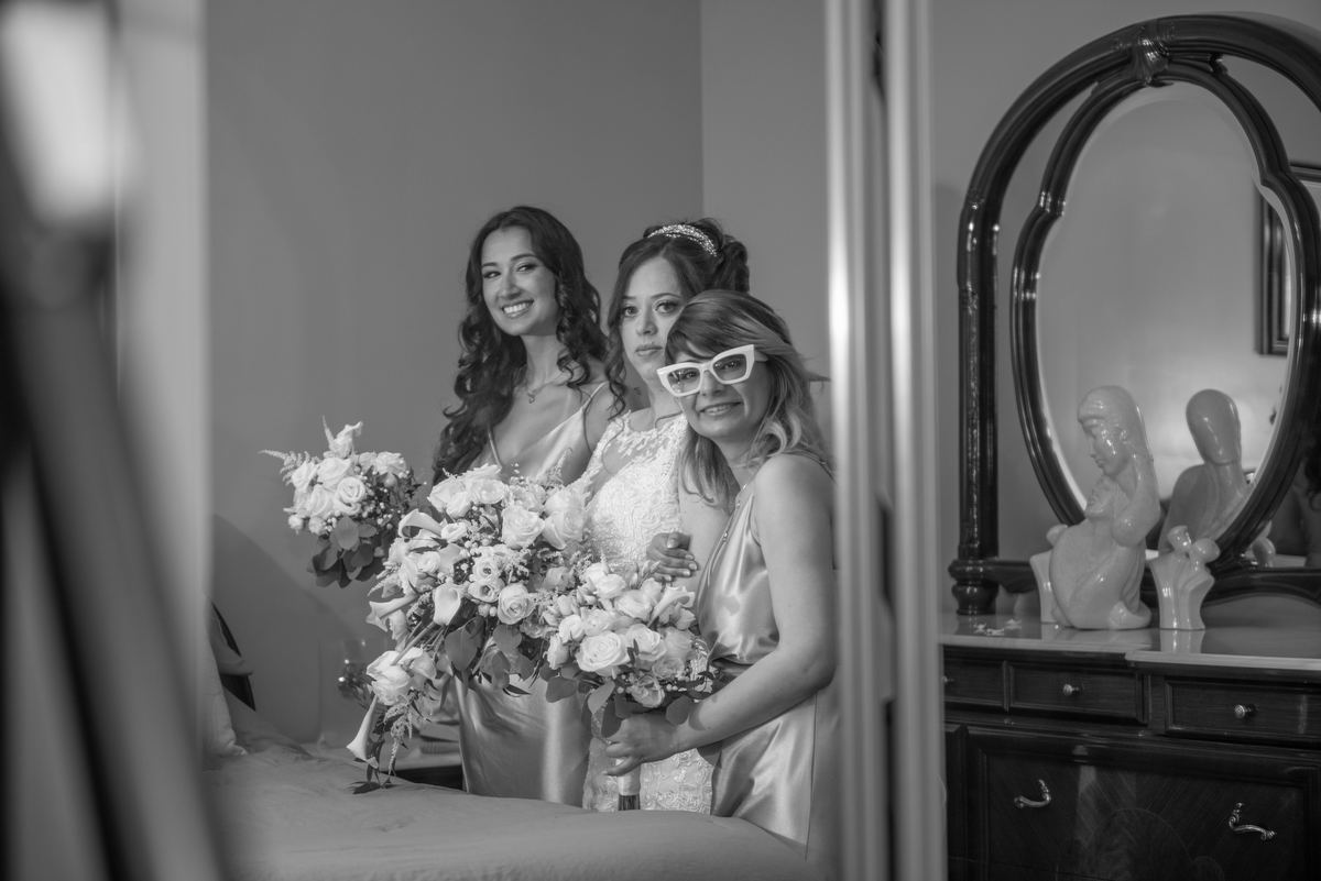 A black-and-white photograph captures three women in a bedroom, smiling at the camera while holding large bridal bouquets.
