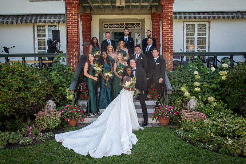 A bride in a white gown and her bridal party pose outdoors on the steps of a brick house surrounded by greenery.