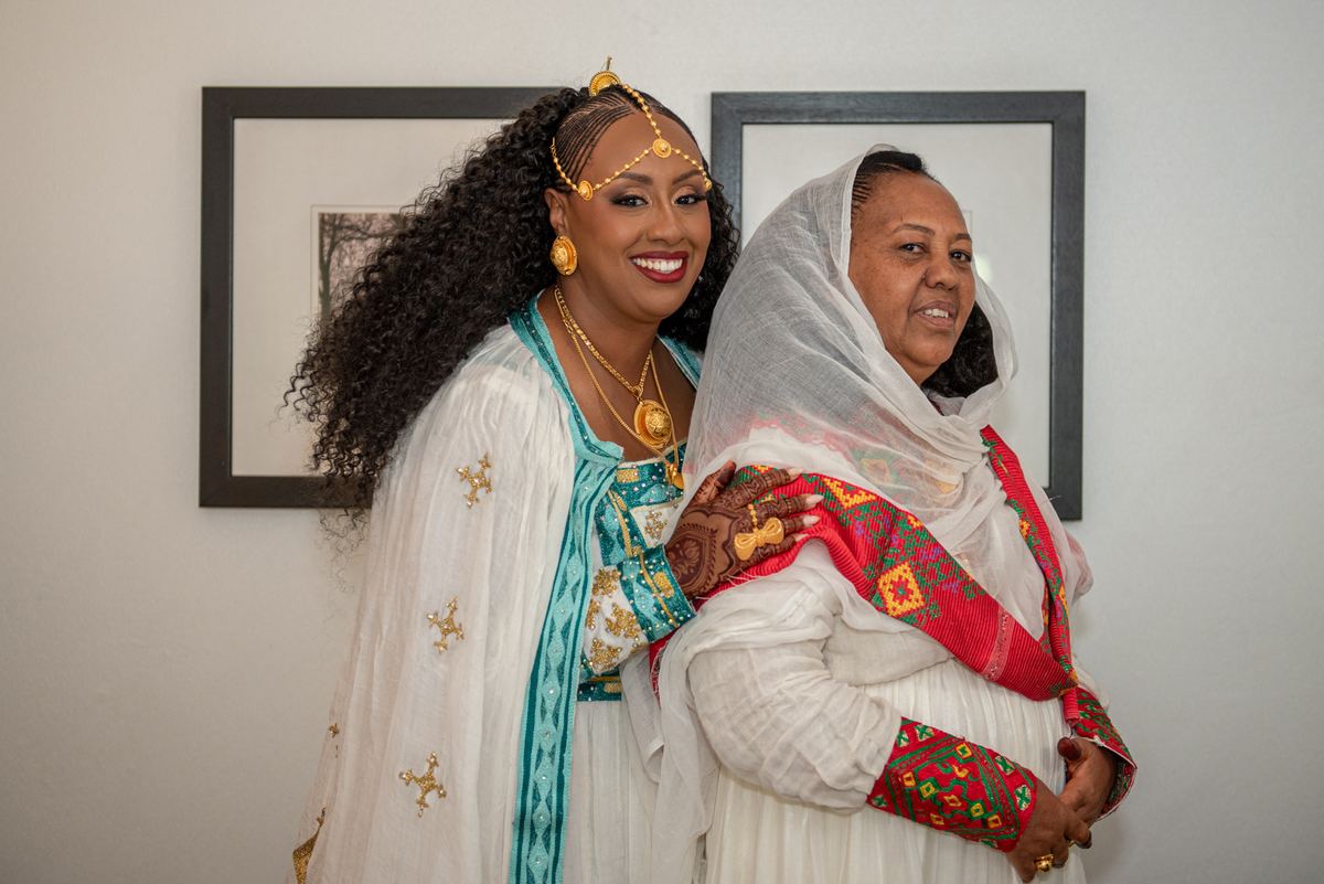Two women in traditional Ethiopian attire embrace warmly, one wearing white and teal with gold jewelry.