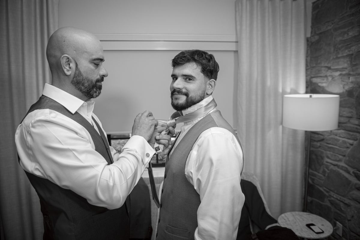 Two men in formal attire, one adjusting the other's tie, illuminated by a lamp casting shadows on a stone wall and curtains.
