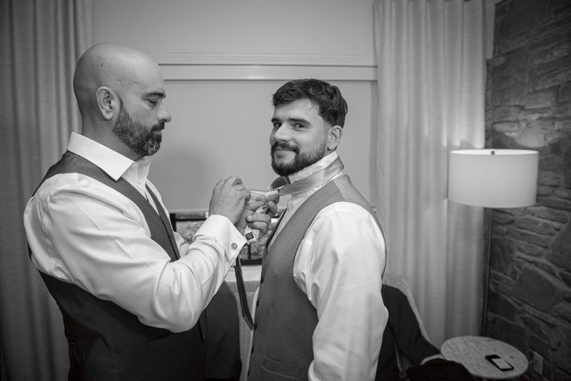 Two men in formal attire, one adjusting the other's tie, illuminated by a lamp casting shadows on a stone wall and curtains.