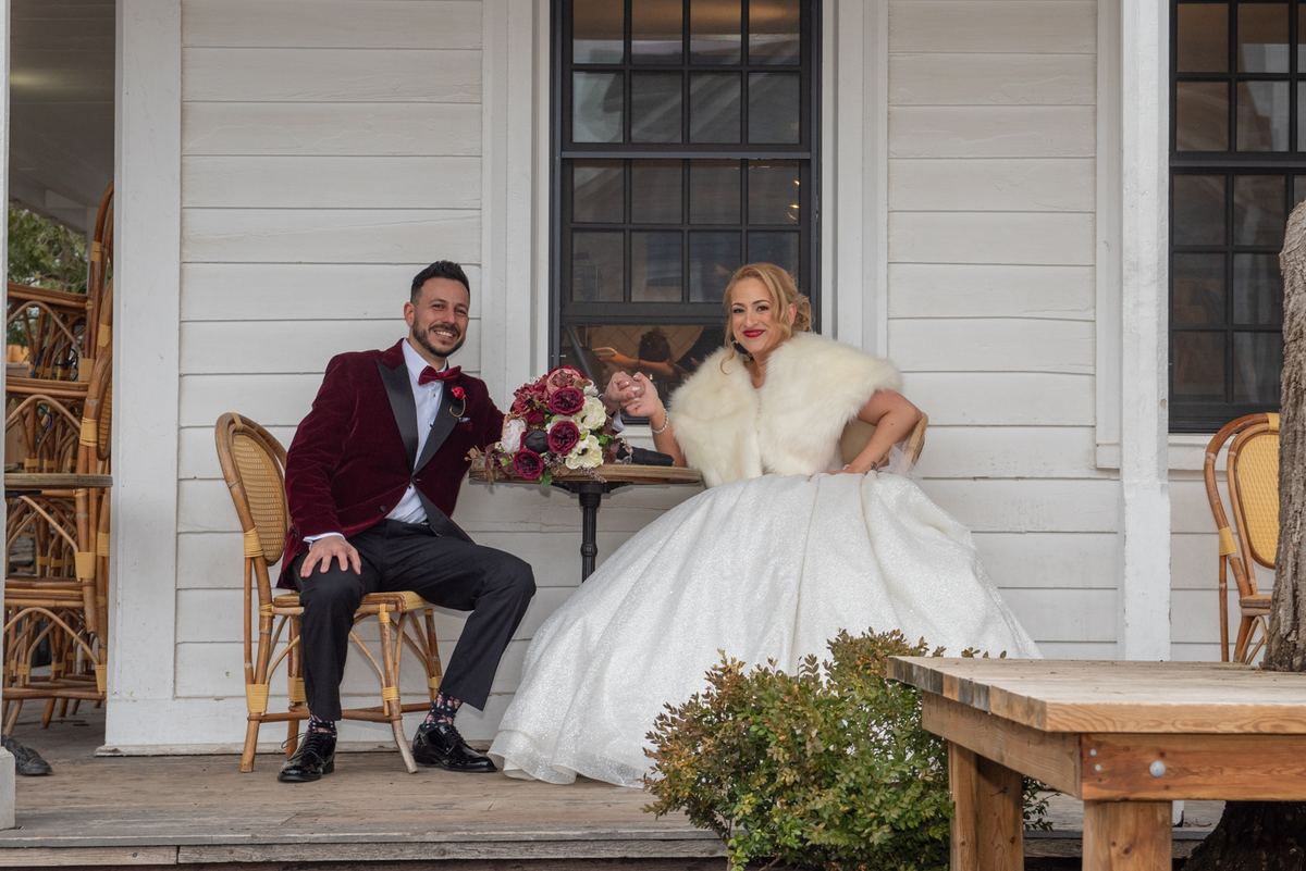 A smiling bride and groom in formal attire sit on a porch with a white clapboard building behind them.