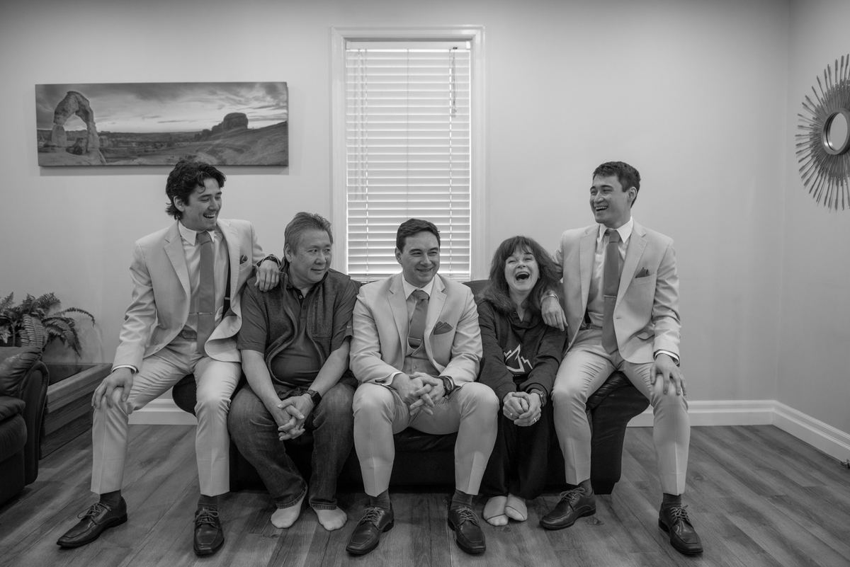 Five people laughing together on a couch in a warmly lit room, illuminated by natural light from a window.