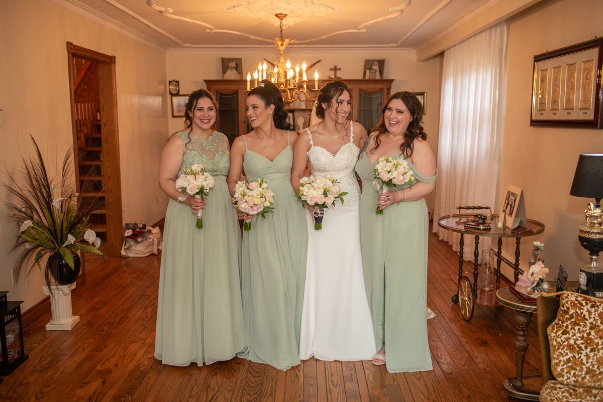 A bride in a white gown and three bridesmaids in sage green dresses smile with bouquets, standing in an ornate home interior.