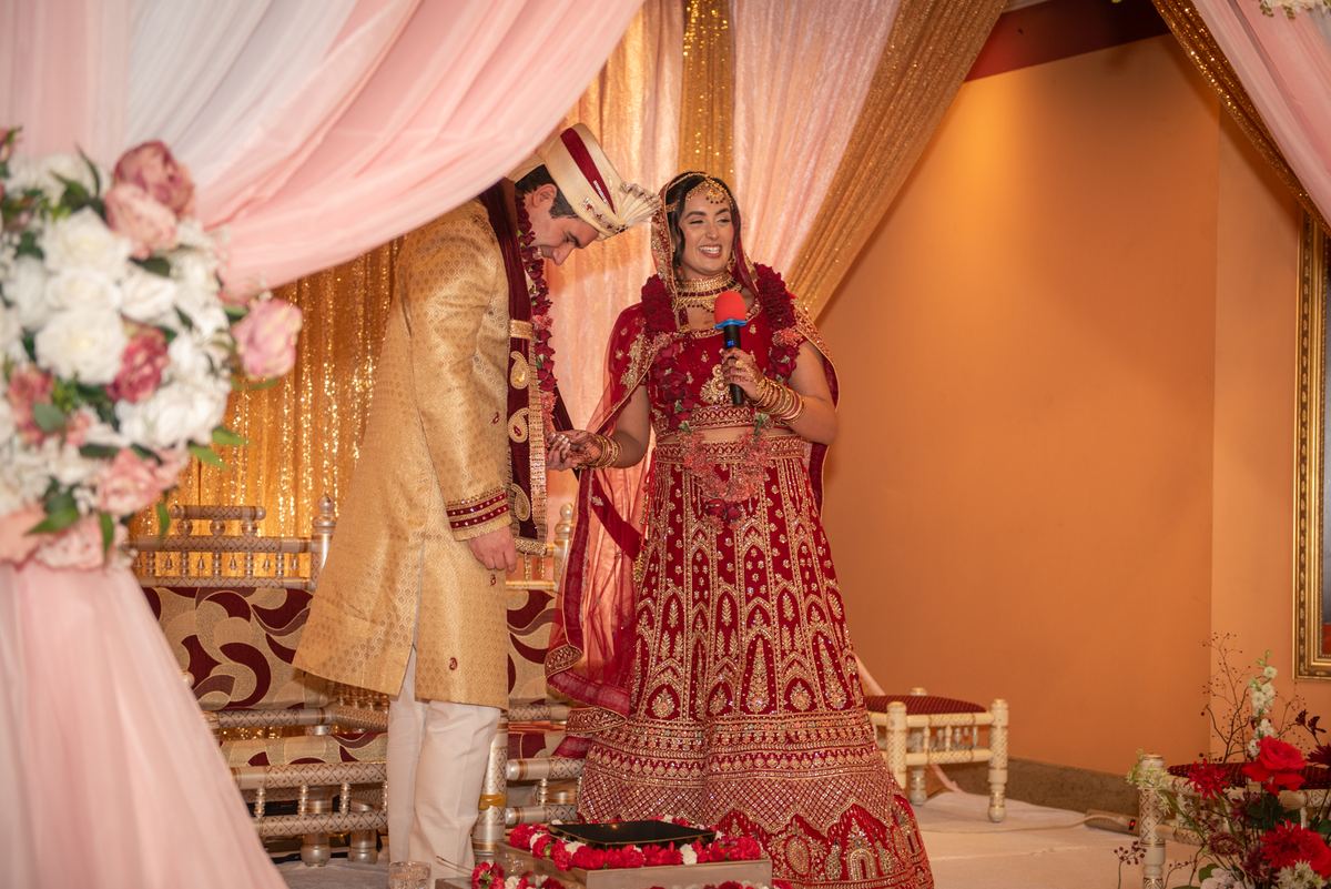 A bride in a red lehenga and her groom in a golden sherwani smile on a decorated stage during their wedding.