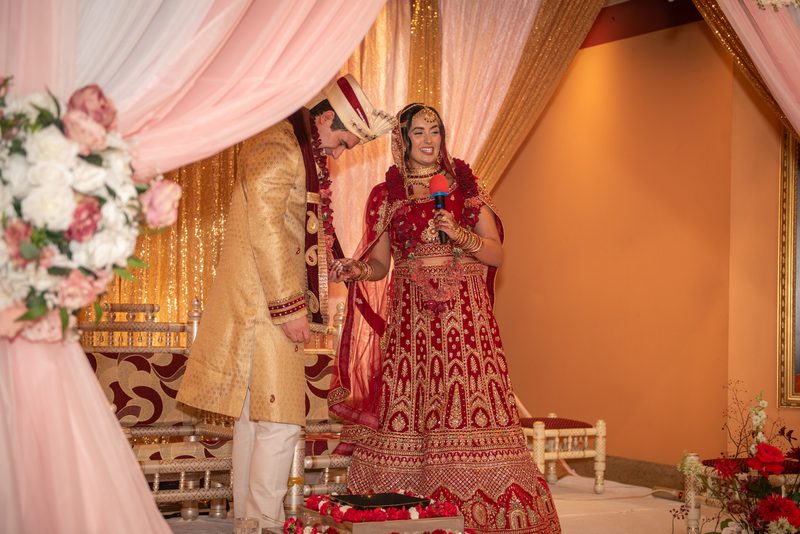 A bride in a red lehenga and her groom in a golden sherwani smile on a decorated stage during their wedding.
