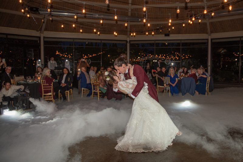A bride and groom hold each other in a first dance surrounded by low white smoke, under warm Edison bulbs strung overhead.