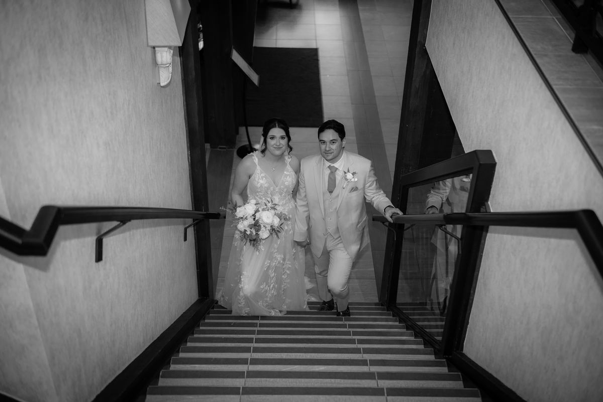 A bride and groom ascend a staircase together in black-and-white, highlighting their attire and the soft indoor lighting.