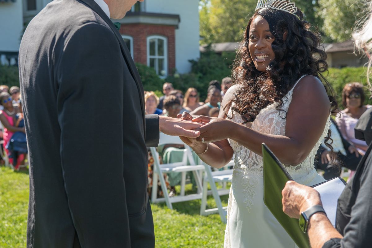 A bride smiles as she places a ring on her groom's finger during an outdoor ceremony, bathed in sunlight.