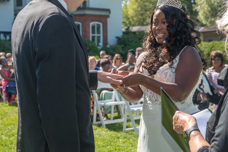 A bride smiles as she places a ring on her groom's finger during an outdoor ceremony, bathed in sunlight.
