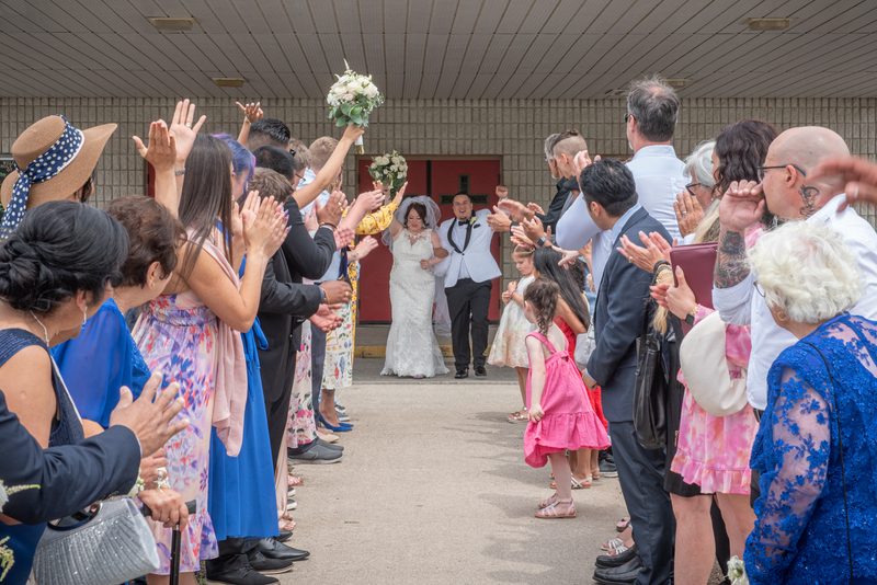 Bride and groom walk down a path flanked by cheering guests, holding her bouquet, in bright daylight.