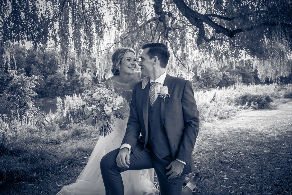 A bride and groom smile under a weeping willow during a wedding photoshoot, in cool blue monochrome.