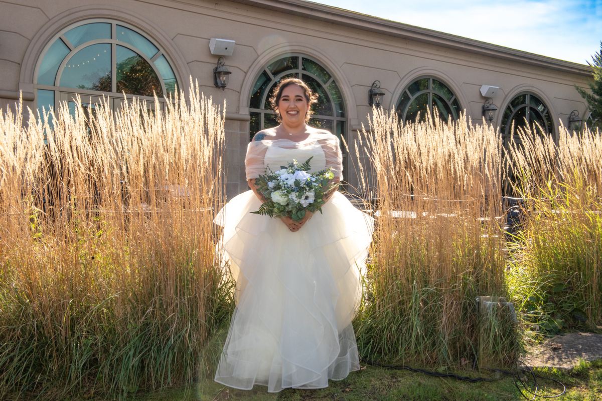 A smiling bride stands in a sunlit outdoor setting, surrounded by tall golden grasses and an arched window building.