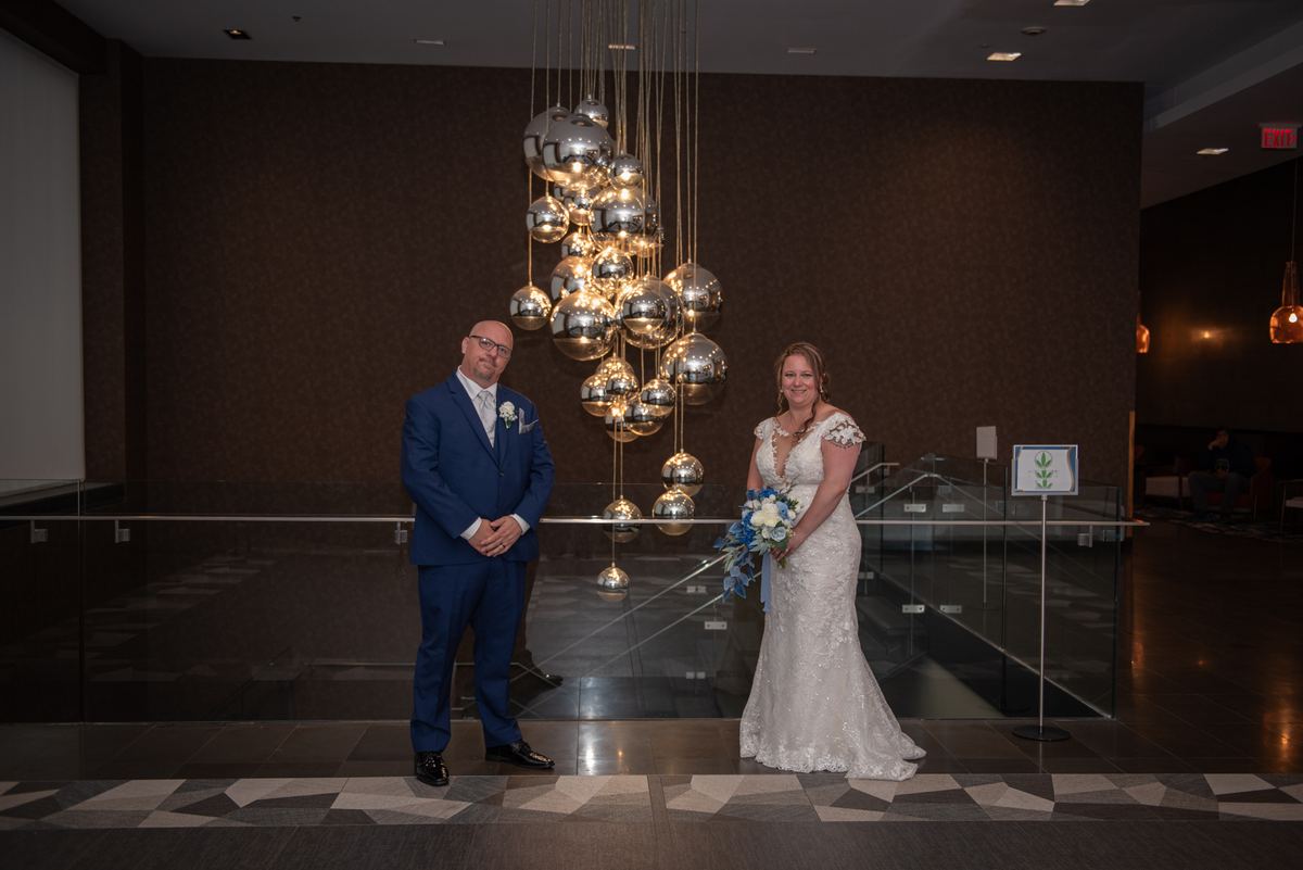 A bride and groom stand in a modern hotel lobby, smiling near a dramatic chandelier of reflective orbs.