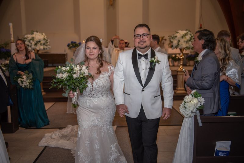 A bride in a lace mermaid gown and veil walks down the aisle with her groom in a white tuxedo toward the altar.