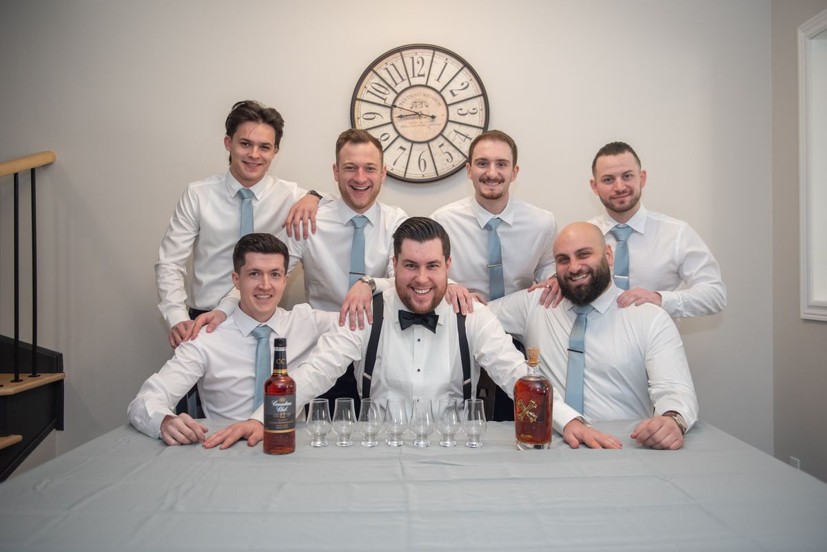 Seven men in white shirts and ties gather around a table with whiskey bottles and glasses, celebrating.