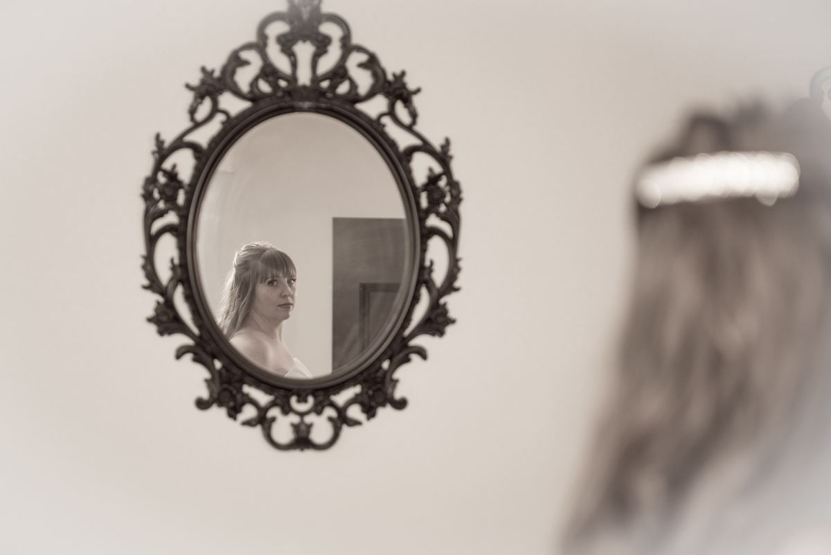 A bride looks into an ornate mirror, her reflection and the silhouette of her veil in soft focus, preparing for her wedding.