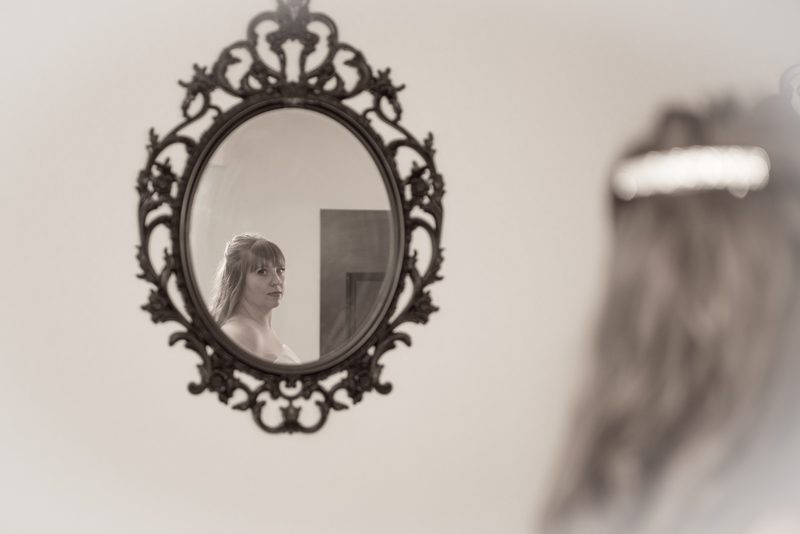 A bride looks into an ornate mirror, her reflection and the silhouette of her veil in soft focus, preparing for her wedding.