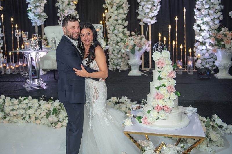 A smiling bride and groom embrace next to their elaborate floral wedding cake surrounded by romantic decor.