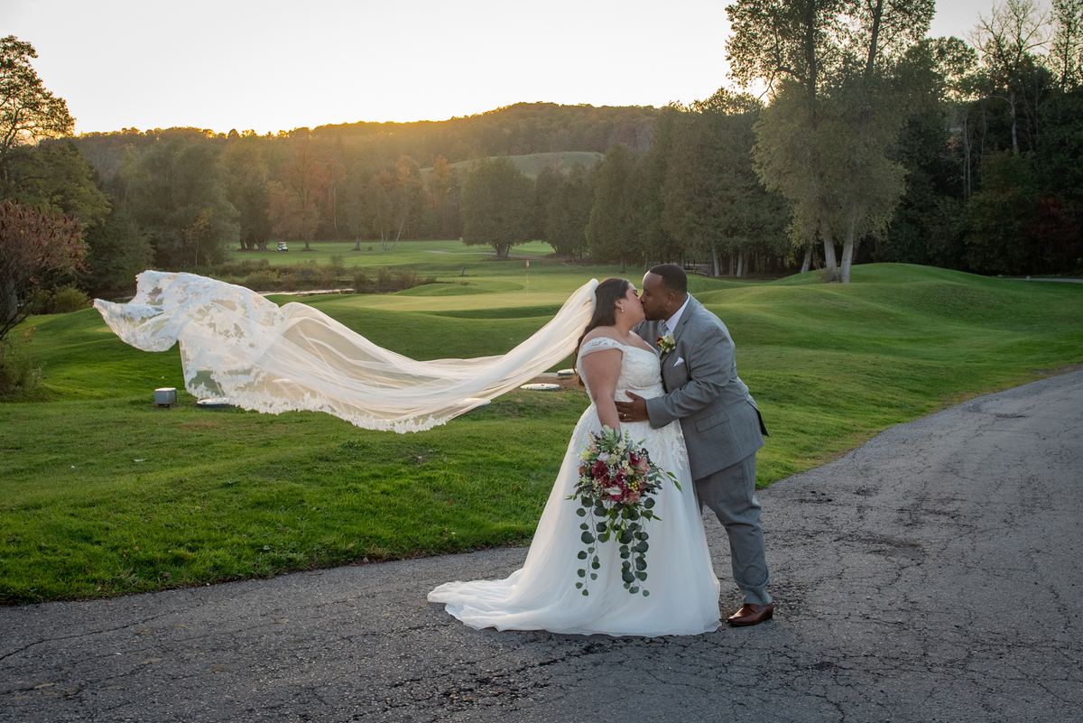 A bride and groom kiss beside a golf course path as the sun sets, their long shadows stretching across the green landscape.