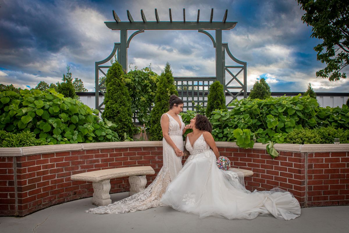 Two brides sharing an intimate moment outdoors, touching faces tenderly beside a brick wall in a garden setting.