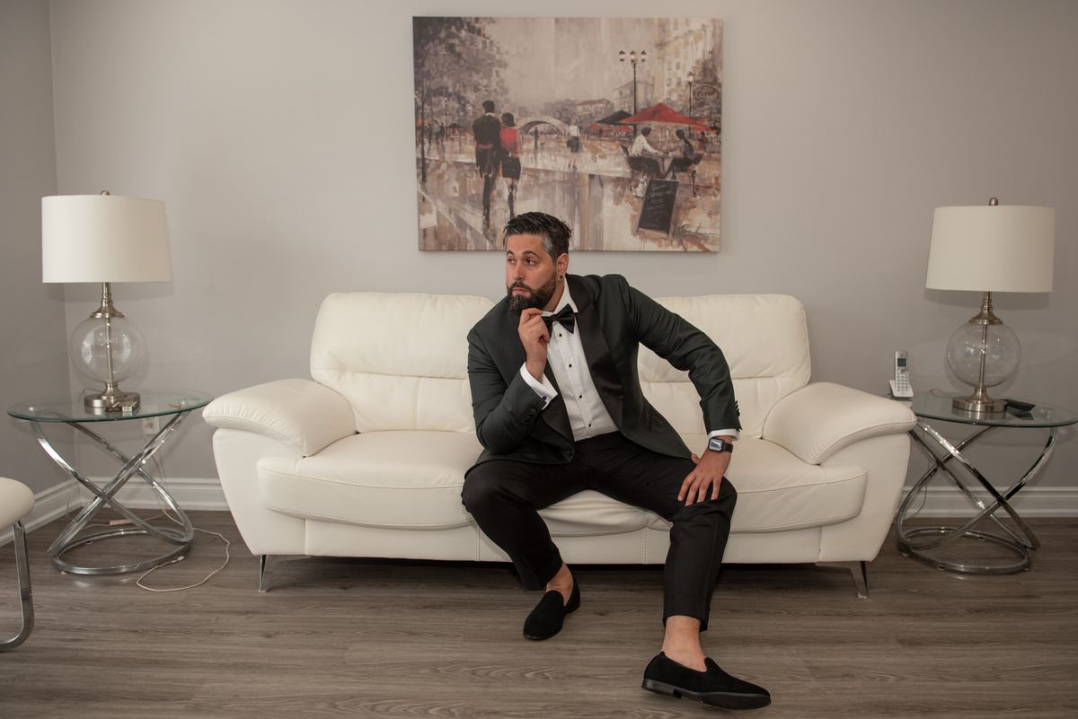 A man in a tuxedo sits thoughtfully on a white sofa in an elegantly lit room, hand resting on his chin.