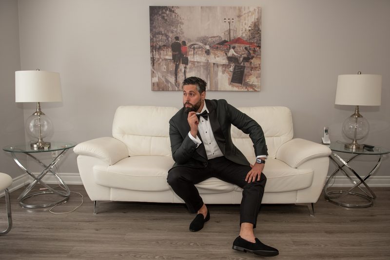 A man in a tuxedo sits thoughtfully on a white sofa in an elegantly lit room, hand resting on his chin.