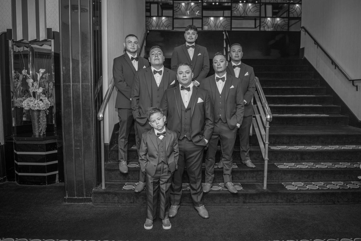 A groom and his groomsmen, including a young boy, pose formally in matching tuxedos on an elegant staircase.