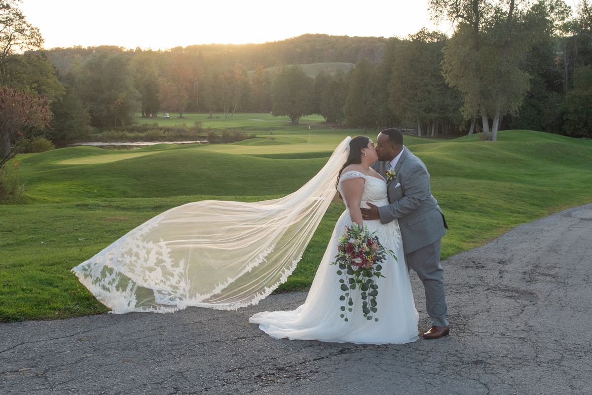 A bride and groom kiss beside a golf course at sunset, their formal attire and the lush greens highlighted by golden light.