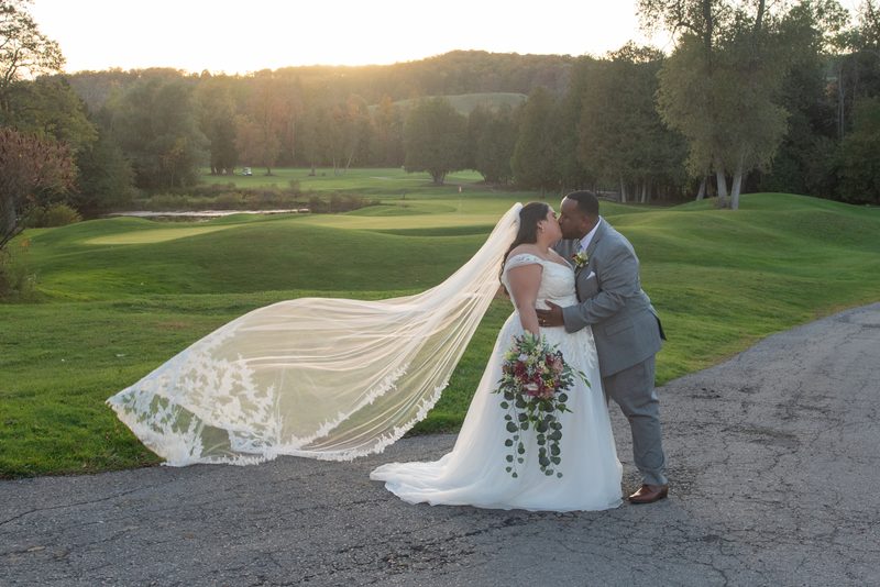 A bride and groom kiss beside a golf course at sunset, their formal attire and the lush greens highlighted by golden light.
