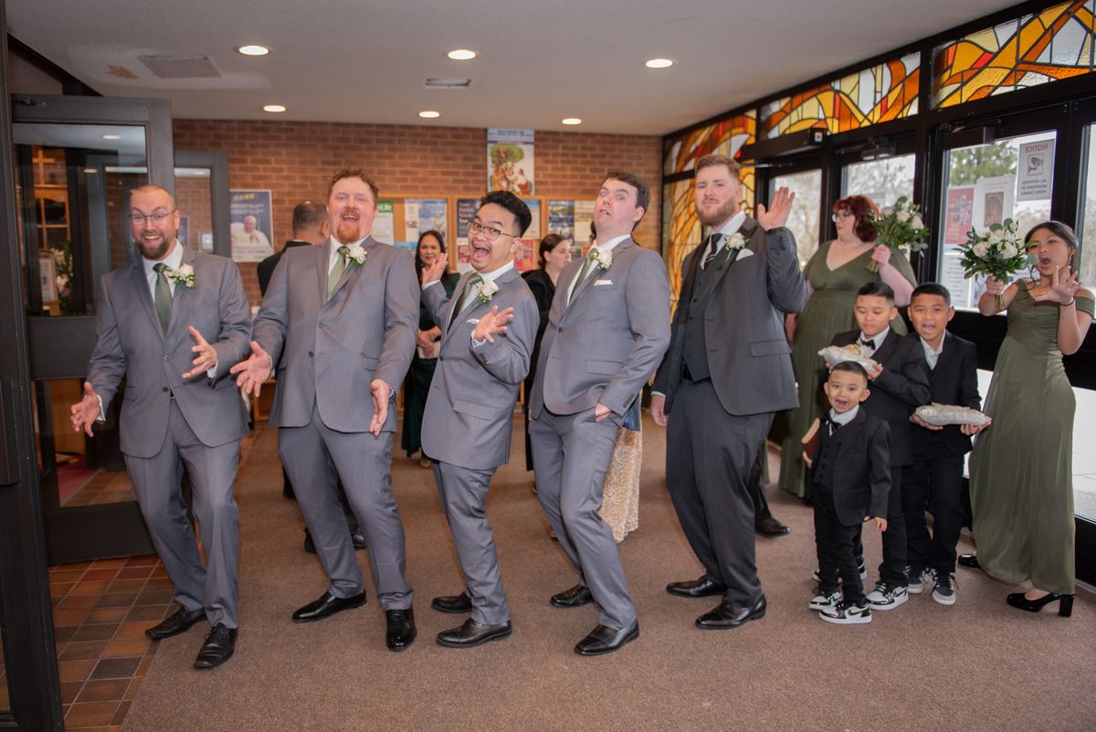 A wedding party in gray suits and young boys in formal wear stand playfully posing indoors with stained glass windows.
