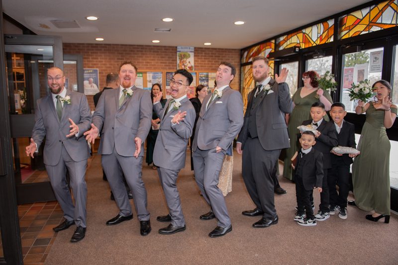 A wedding party in gray suits and young boys in formal wear stand playfully posing indoors with stained glass windows.