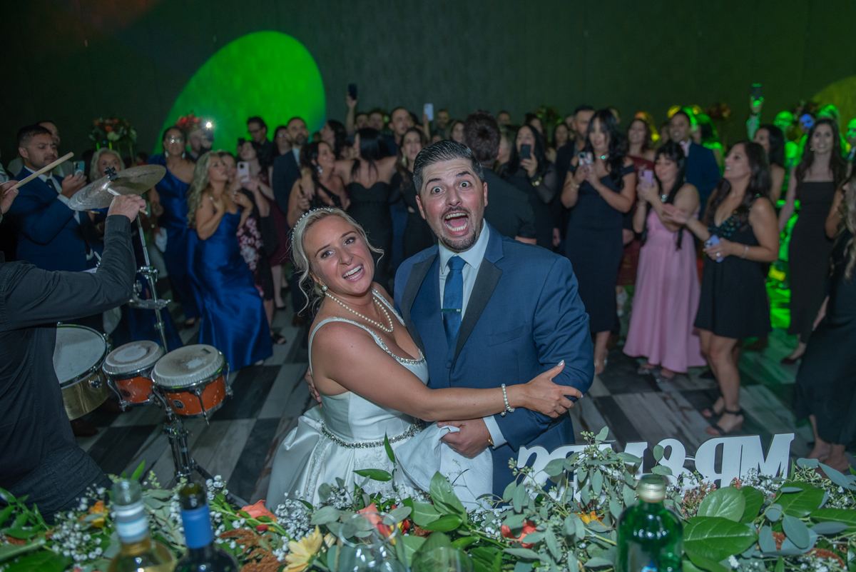 Bride and groom smile and embrace at celebration, surrounded by cheering guests and a live band under green stage lights.