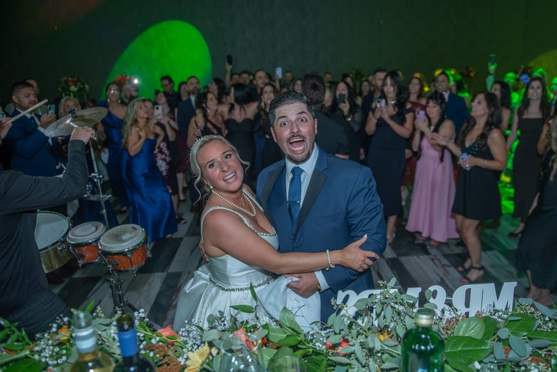 Bride and groom smile and embrace at celebration, surrounded by cheering guests and a live band under green stage lights.