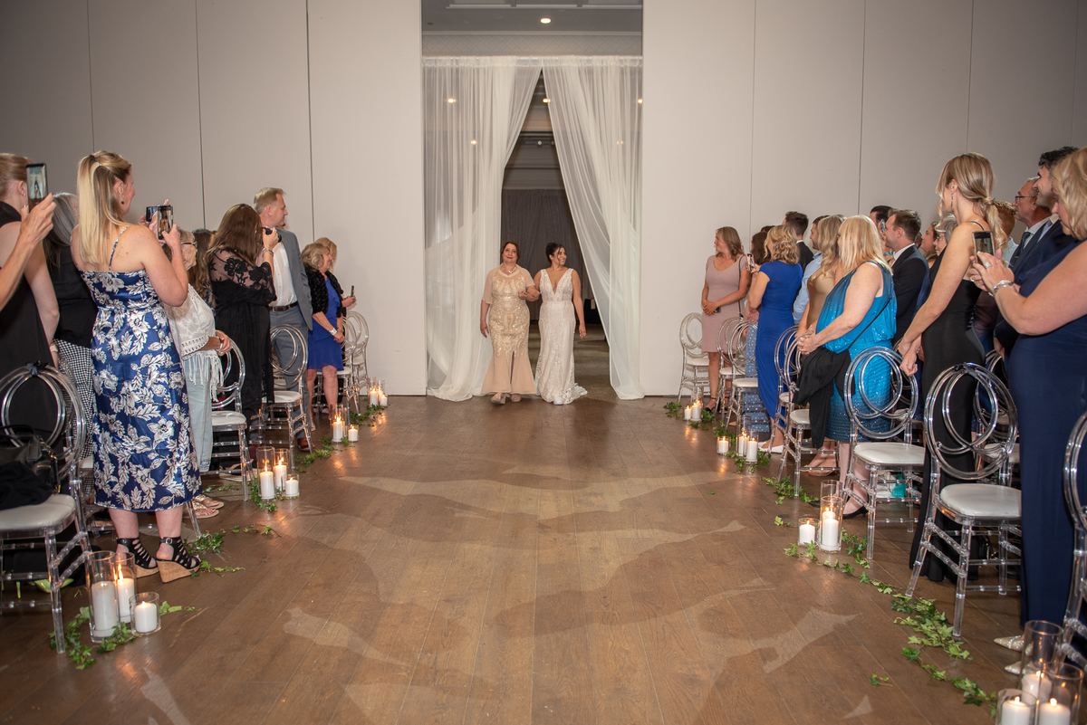 A bride in a white gown walks down a candlelit aisle toward the altar, escorted by a family member in an elegant gold dress.