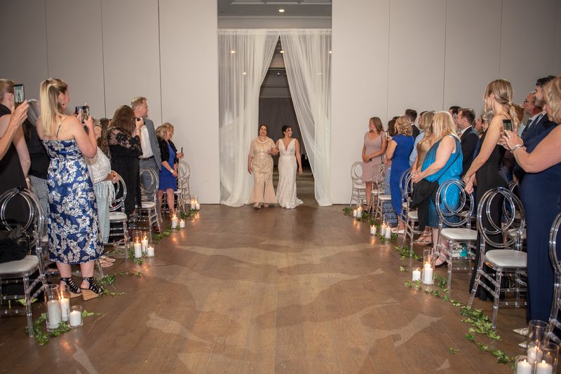 A bride in a white gown walks down a candlelit aisle toward the altar, escorted by a family member in an elegant gold dress.