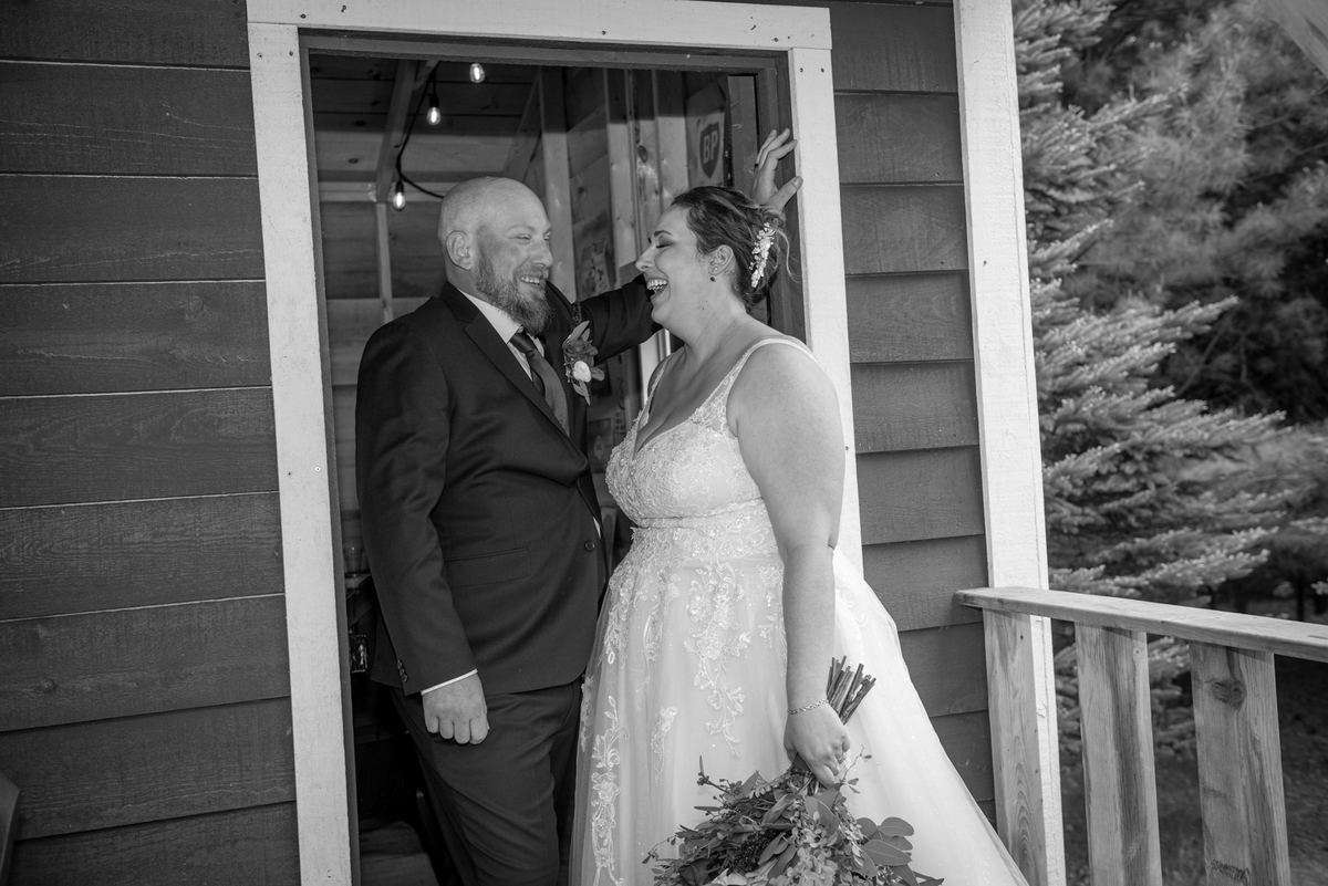 A smiling groom and laughing bride share an intimate moment in a rustic doorway, bathed in soft black-and-white light.