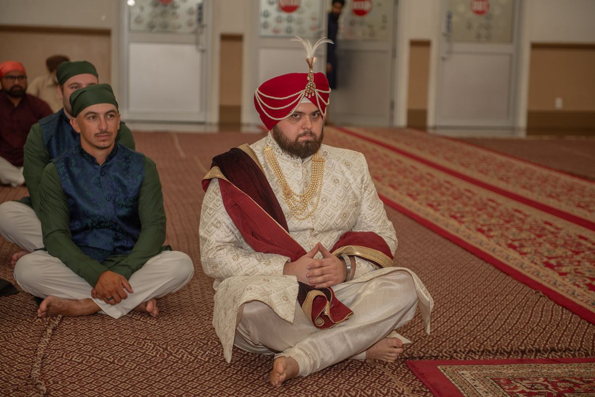 A groom in ceremonial attire sits cross-legged with other men in traditional clothing, suggesting a religious gathering.