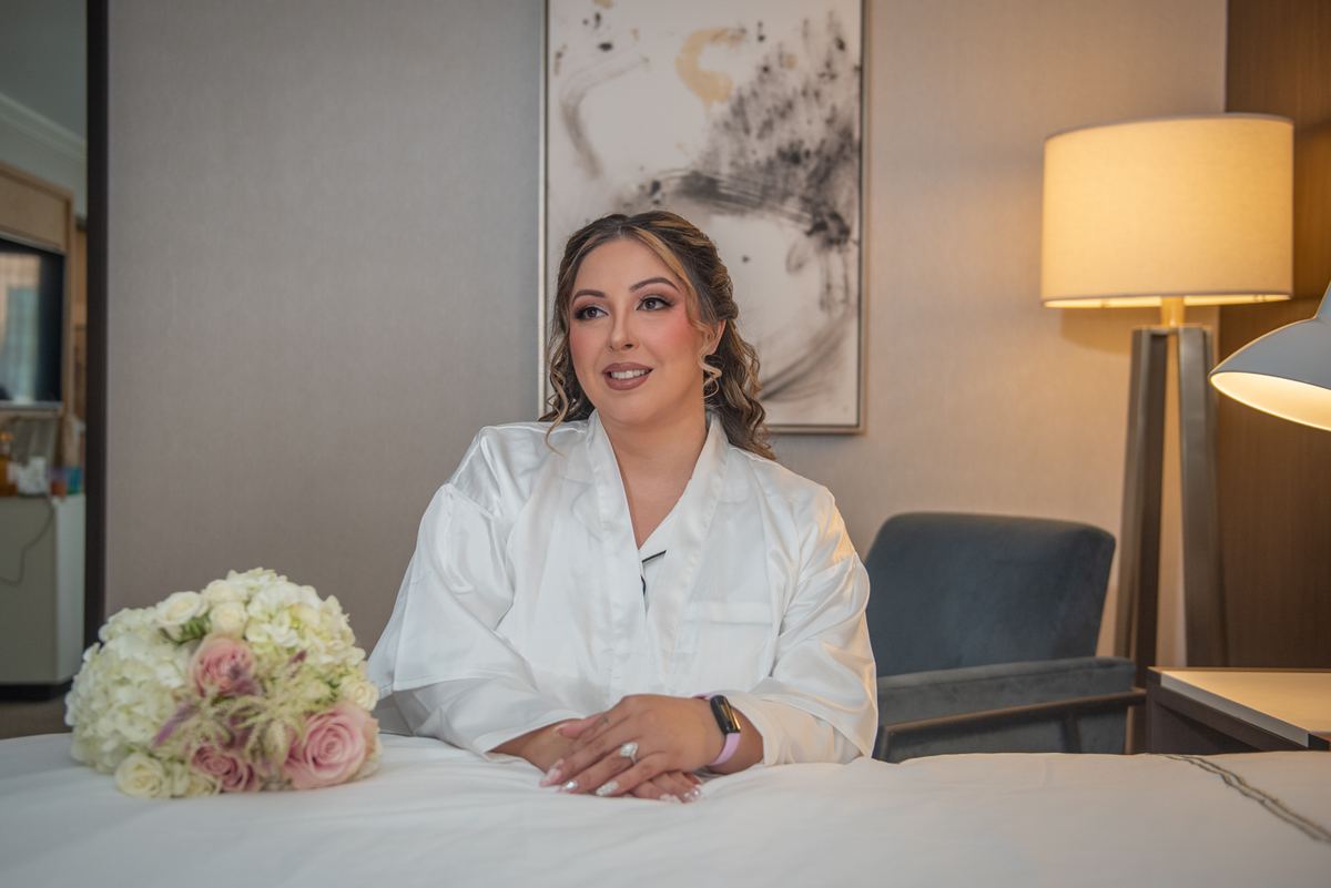 A bride in a white satin robe sits by a bouquet of flowers, smiling softly in the warm glow of a floor lamp.