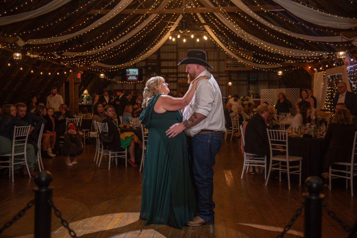 A couple dances in a barn decorated with string lights and fabric, he in a cowboy hat, she in a green gown.