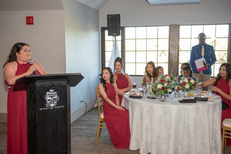 A woman in a red gown speaks at a podium while guests listen, surrounded by natural light and matching red dresses.