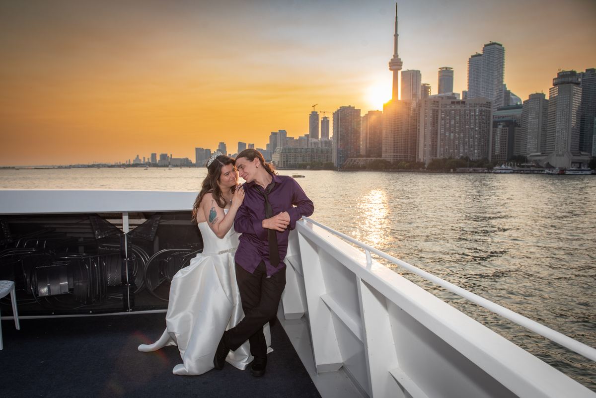 A bride and groom embrace on a ferry deck at sunset, with the Toronto skyline glowing in the background.