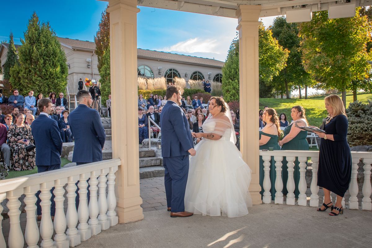 A bride and groom stand under an outdoor pavilion during their wedding ceremony, guests seated behind them.